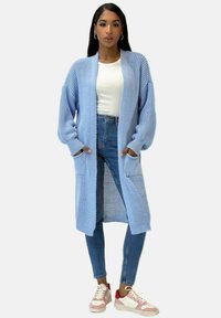 Cardigan lavorato a maglia di colore azzurro chiaro, dal taglio morbido, con maniche lunghe e due tasche frontali, indossato sopra un top bianco aderente e jeans in denim.