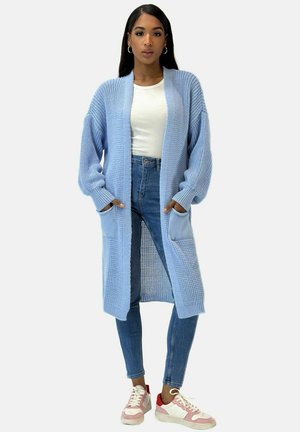 Cardigan lavorato a maglia di colore azzurro chiaro, dal taglio morbido, con maniche lunghe e due tasche frontali, indossato sopra un top bianco aderente e jeans in denim.