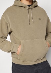 Felpa beige con tasca frontale e stringhe indossata da una persona con le mani nella tasca, abbinata a pantaloni scuri.