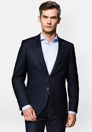 Granatowy, dopasowany garnitur z klapami typu notch i dwoma guzikami, zestawiony z jasnoniebieską koszulą oxford. Gładki materiał o minimalistycznym wzornictwie.