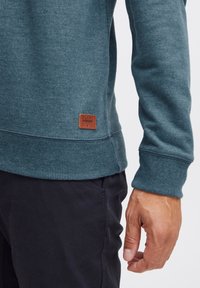 Sudadera de color azul verdoso con puños y dobladillo acanalados, que presenta un parche de logo de cuero marrón. Tejido de mezcla de algodón, textura suave, diseño ajustado.