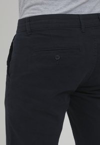 Pantaloni navy scuri in cotone con una consistenza liscia, dotati di una sola tasca posteriore con chiusura a bottone e passanti per la cintura.