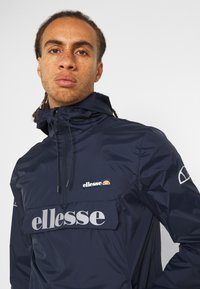 Tengerkék széldzseki elülső zsebbel, rugalmas mandzsettákkal és állítható kapucnival. Az "ellesse" logó szürke színben található a mellkasán.