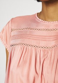 Blusa rosa realizzata in tessuto morbido, con scollatura rotonda, dettagli in pizzo intricati e texture arricciata sotto il petto.
