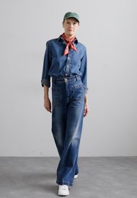 Denim shirt and high-waisted flared jeans in dark blue, with a red bandana and green cap. White sneakers complete the outfit.