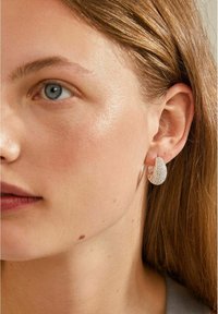 Pendiente de aro de plata con una superficie texturizada y brillante, acompañado de un primer plano de la cara de un modelo que muestra su tono de piel natural y cabello.