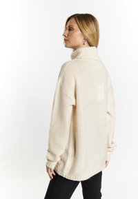 Beige, oversized pulóver magas gallérral, texturált kötésű, bordás mandzsettával, fekete nadrágra stílusosan kombinálva. Puha anyag, laza szabással.