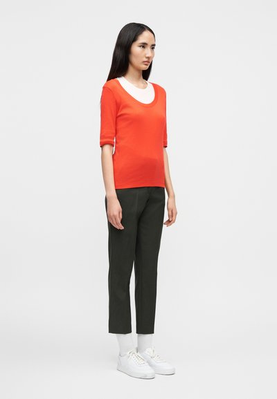 Γυναίκα με μακριά σκούρα μαλλιά που φοράει πορτοκαλί μπλουζάκι με κοντά μανίκια πάνω από λευκή μπλούζα, σκουρόχρωμο παντελόνι μέχρι τον αστράγαλο, λευκές κάλτσες και λευκά αθλητικά παπούτσια.