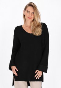 Pulover negru supradimensionat cu decolteu în V, mâneci lungi, textură cu striații și fante laterale, purtat cu pantaloni de culoare deschisă. Design simplu și modern.