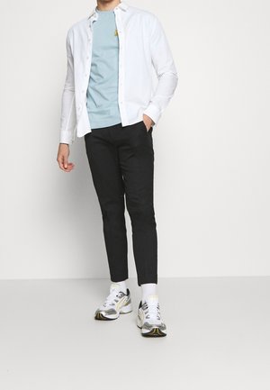 Chemise blanche boutonnée sur un t-shirt bleu clair avec un logo jaune, accompagnée d'un pantalon slim noir et de baskets argentées.