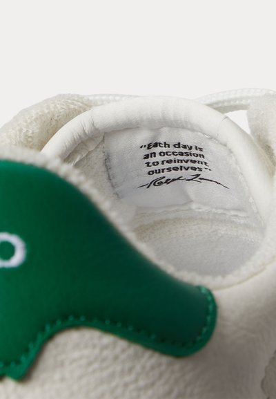 Biela päta tenisky so zeleným koženým štítkom, na ktorom je látkový štítok s citátom: „Každý deň je príležitosťou na obnovu seba samého.“