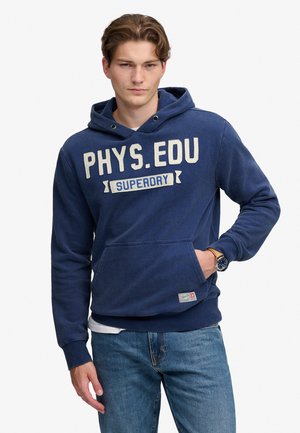 Ung mand iført en blå hættetrøje med teksten "PHYS.EDU SUPERDRY" og blå jeans, poserer med den ene hånd i hættetrøje-lommen.