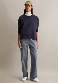 Granatowa bluza z okrągłym dekoltem i ściągaczami przy rękawach, w zestawieniu z jasnoszarymi spodniami dresowymi. Na głowie beżowa czapka z daszkiem z wyszytym numerem "16".