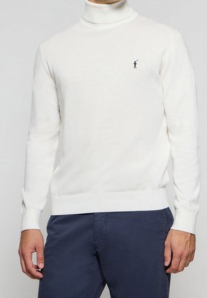 Mann trägt einen weißen Rollkragenpullover mit kleinem marineblauem Logo auf der Brust und dunkelblaue Hose, steht vor einem schlichten Hintergrund.