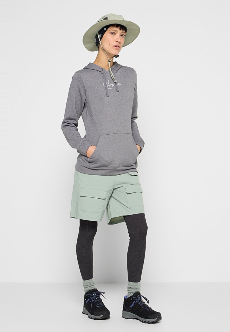 Persona que lleva una sudadera gris, pantalones cortos de carga verde claro, mallas negras, botas de senderismo y un sombrero de ala ancha con correa para el mentón, apoyada contra una pared blanca.