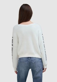 Weißer flauschiger Pullover mit schwarzem Text auf den Ärmeln, kombiniert mit blauen Jeans. Der Pullover hat einen weiten Halsausschnitt und eine lockere Passform.