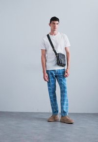 Bílé tričko, kalhoty s modrým vzorem, béžové boty a malá černá kabelka přes tělo se ramenním popruhem. Jednoduché šedé pozadí.