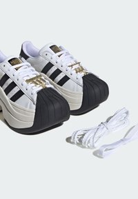 Λευκά platform sneakers Adidas με μαύρες προστατευτικές μύτες, μαύρες ρίγες, χρυσές ετικέτες κορδονιών και επιπλέον λευκά κορδόνια σε ανοιχτό φόντο.