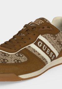 Καφέ και μπεζ αθλητικό παπούτσι Guess με μοτίβο λογότυπου, λευκές ρίγες και καφέ κορδόνια, που δείχνει λεπτομέρειες από πλευρά και εμπρός.