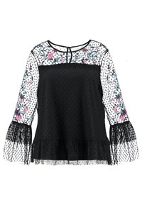 Blouse noire à motif à pois, avec des manches longues transparentes ornées de broderies florales et un ourlet à volants. Tissu doux avec une légère texture.