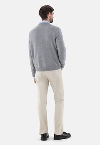 Grauer gestrickter Pullover mit geripptem Saum, kombiniert mit hellbeigen Hosen. Die Person steht in schwarzen Schuhen, von hinten sichtbar vor einem weißen Hintergrund.