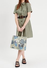 Sac en toile avec un motif floral bleu, poignées renforcées et finition contrastante. Porté avec une robe verte à boutons et des sandales noires.