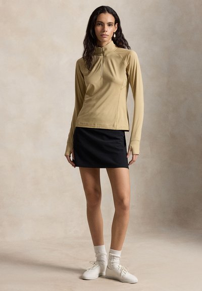 Γυναίκα που φοράει μπεζ μακρυμάνικο μπλουζάκι με φερμουάρ, μαύρη μίνι φούστα, λευκές κάλτσες και λευκά παπούτσια με κορδόνια, στέκεται μπροστά σε ουδέτερο φόντο.