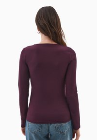 Donna con lunghi capelli castani che indossa una maglietta aderente a maniche lunghe di un viola scuro e jeans blu, vista di spalle su uno sfondo bianco.