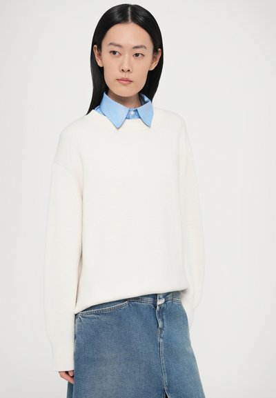 Fehér kötött pulóver lejött vállakkal, világoskék gombos ing fölött viselve, kék farmer szoknyával párosítva. Simább textúra.