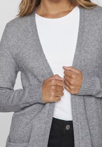 Cardigan gris en tricot de fil doux, offrant une coupe décontractée, une ouverture devant, des poignets côtelés et deux poches avant. Porté sur un haut blanc.