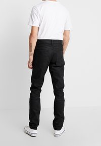 Jeans negros de corte ajustado con bolsillos traseros estándar, combinados con una camiseta blanca y zapatillas blancas. Textura suave y diseño simple.