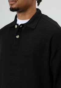 Zwart gebreid poloshirt met een kraag en twee beige knopen. De stof heeft een gestructureerde uitstraling.