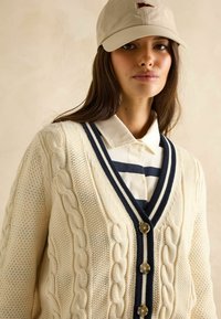 Kreemjas kudumiga kardigan, millel on kaabelmustritega detailid, tumesinised aktsendid ja nööbid. Kantud valge särk üle, millel on tumesinised triibud. Beež müts väikese punase embleemiga.