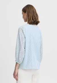 Blusa azzurro chiaro con motivo a onde testurizzato, maniche trasparenti e polsini elastici. Collo rotondo e design sul retro con dettagli sottili.