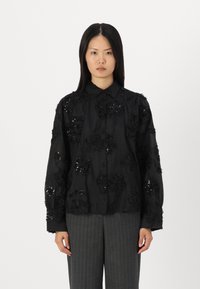 Camisa preta de botão com bordados florais e acentos de lantejoulas. Mangas longas, corte descontraído, feita de tecido suave. Combinada com calças cinzas listadas.