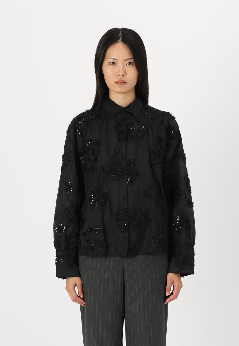 Camisa preta de botão com bordados florais e acentos de lantejoulas. Mangas longas, corte descontraído, feita de tecido suave. Combinada com calças cinzas listadas.