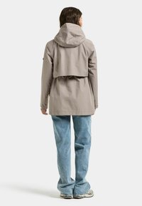 Frau trägt eine beige Kapuzen-Regenjacke, hellblaue Jeans und Turnschuhe, steht mit dem Rücken zum Betrachter vor einem einfarbigen Hintergrund.