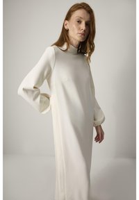 Witte jurk met lange mouwen, hoge hals en pofmouwen. Glad materiaal, tot op de knie, met subtiele naaddetails.