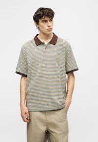 Νέος άνδρας που φοράει μπλούζα πόλο με μπεζ ρίγες, καφέ γιακά και μανσέτες, συνδυασμένη με ανοιχτό χακί παντελόνι, στέκεται μπροστά σε απλό φόντο.