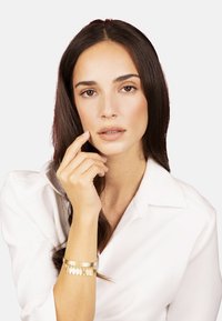 Pulsera de oro con acentos colgantes, acabado liso y brillante. La modelo lleva una camisa blanca, tiene el cabello largo y oscuro, y sostiene un dedo en sus labios.