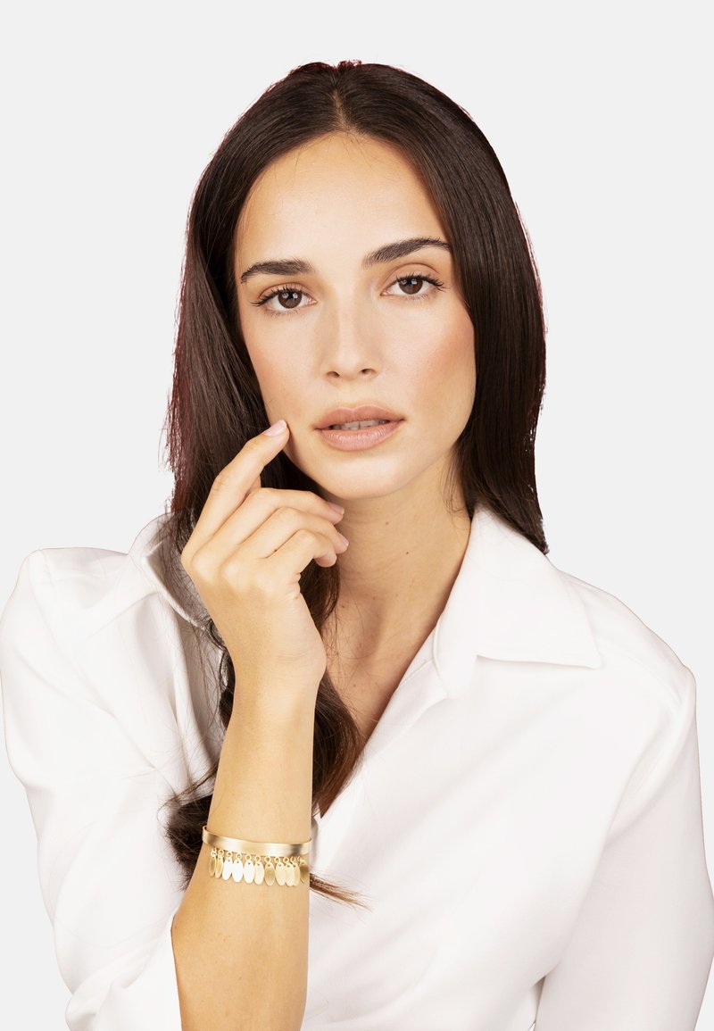 Pulsera de oro con acentos colgantes, acabado liso y brillante. La modelo lleva una camisa blanca, tiene el cabello largo y oscuro, y sostiene un dedo en sus labios.