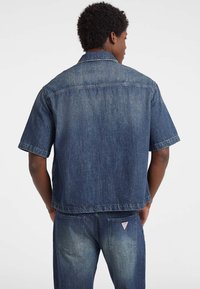 Giubbotto in denim blu scuro, maniche corte, vestibilità ampia, cuciture visibili e un'etichetta sul retro. Design pulito con una finitura testurizzata.