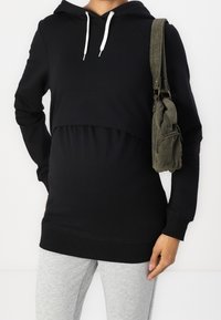 Svart huvtröja med en kortad design och vita dragsnören. Kombinerad med grå sweatpants och en grön snörväska.