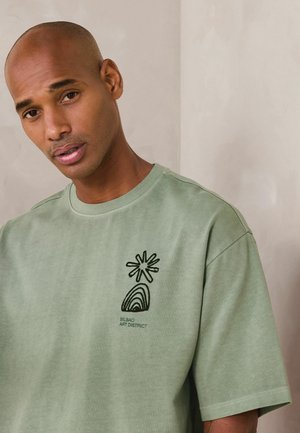 Mann mit kahl rasiertem Kopf trägt ein hellgrünes T-Shirt mit schwarzem Bilbao Art District Grafik auf der Brust, steht vor einer schlichten beigen Wand.