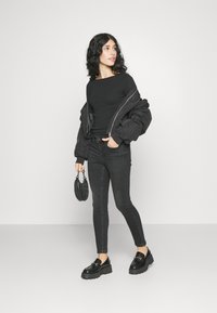 Μαύρη βομβιστική ζακέτα πάνω από μαύρο τοπ, συνδυασμένη με στενές μαύρες τζιν παντελόνες και χοντρές μαύρες πλατφόρμες. Κρατάει μία μικρή μαύρη τσάντα.