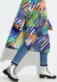 Impermeabile multicolore con motivo geometrico, abbinato a jeans blu e stivali combat argentati con lacci. Presenta una vestibilità comoda e dettagli con bottoni.