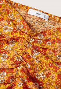 Haut floral jaune avec un col smocké, orné de fleurs rouges, oranges et blanches sur un tissu doux. L'étiquette indique "MNG."