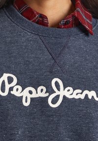 Felpa blu navy con finitura testurizzata, caratterizzata dal logo "Pepe Jeans" ricamato in bianco sul petto, indossata sopra una camicia a quadri rossa.