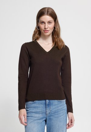 Jeune femme aux cheveux bruns mi-longs portant des boucles d'oreilles anneaux argentés, un pull col en V marron foncé et un jean bleu clair, debout.