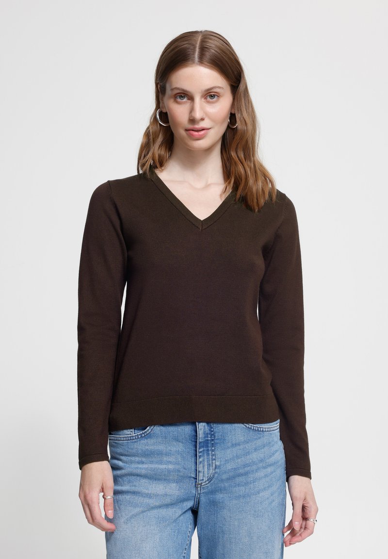 Jeune femme aux cheveux bruns mi-longs portant des boucles d'oreilles anneaux argentés, un pull col en V marron foncé et un jean bleu clair, debout.